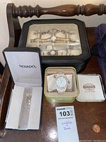 Photo showing multiple ladies watches in a black glass-topped display box, a Fossil watch inside an open green tin box, and an empty Movado jewelry box on a wooden surface.