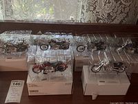 Six Harley-Davidson replica motorcycle models displayed in clear plastic protective cases on white display bases arranged on a table with lace curtain background.