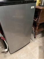 Closed Magic Chef mini fridge showing silver door and black top next to wooden side table.