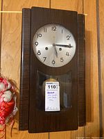 Full front view of mini grand wall clock with wooden case on wood floor background.