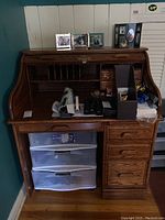 Front view of the oak roll top desk with various office supplies inside the open roll top compartment and plastic drawers underneath.