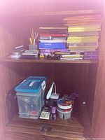 Office supplies on two wooden shelves. Top shelf contains papers, notepads, pens, pencils, calculator, and plastic container. Bottom shelf contains another calculator, tape dispenser, staplers, scissors, and rotary paper clip container.