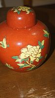 Close-up side profile showing red ceramic surface with hand-painted yellow flowers and green leaves on the jar body and lid.