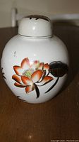 Front angle view of white ceramic vase with orange floral lotus design and removable lid