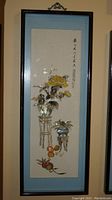 Framed vertical Asian shell art featuring flowers, fruit, and calligraphy.