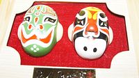 Two painted clay mask figurines mounted on red felt inside a wooden frame.
