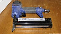 Blue Campbell Hausfeld pneumatic nailer placed on a wooden surface showing the side label and ergonomic grip