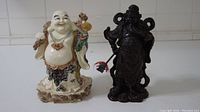 Pair of two Asian statues showing Laughing Buddha and Guan Yu statue side by side on white surface.