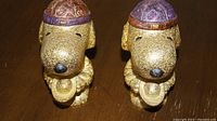 Pair of gold-colored dog-shaped money banks side by side on a wooden surface with purple hats and gold coins in front.