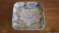 Square decorative plate showing detailed floral pattern with large pink flower and smaller blue flowers, accented with gold background
