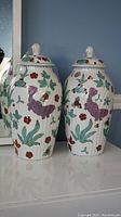 Pair of tall white ceramic lidded jars with purple fish, green leaves, and red flowers, ribbed texture visible