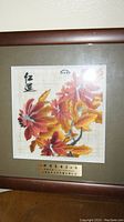 Framed artwork showing red and orange floral scene with Chinese calligraphy in upper left. Mounted on beige mat, square format.