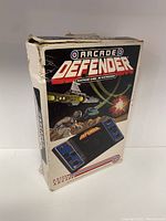 View of the front and side of the Entex Arcade Defender handheld game box showing game branding and vintage sci-fi artwork of spaceships.