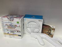 Full view of 12 stacked Wii game cases with title labels facing forward, boxed Wii Wheel controller, and additional Wii game disc outside the stack.