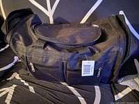 Black nylon duffle bag lying on patterned bedding showing front and top compartments with tags attached.