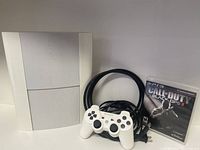 Full view of white PS3 Super Slim console, white DualShock controller, and game case for Call of Duty Black Ops II with cables behind.