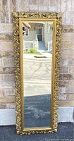 Full front view of the vintage Syroco mirror leaning against brick wall outdoors, showing entire decorative gold tone frame and mirror glass.