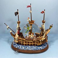 Front view of the Peter Pan pirate ship musical snow globe showing the central glass globe with characters inside and decorative ship details on the base and sides.