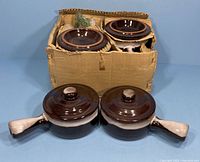 Two ceramic French onion soup bowls with lids and handles placed in front of the original cardboard box holding the remaining bowls with packing material.