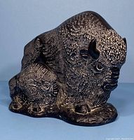 Front and side view of dark soapstone sculpture featuring a bison and calf in textured detail.
