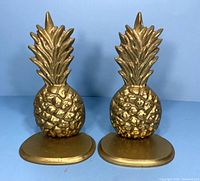 Front view of vintage brass pineapple bookends showing textured pineapple bodies and leafy tops on oval bases.