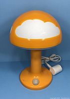 Front angle view of orange mushroom table lamp with white clouds, showing power cord and button on base.