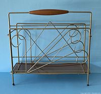 Front view of vintage mid-century modern magazine rack in brass tone metal with wooden handle and laminate shelf.