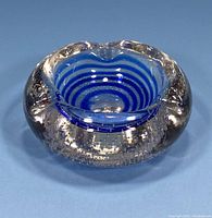 Top angle view of the round Murano glass ashtray showing blue concentric stripes inside thick clear glass with air bubbles, three cigarette rests visible.