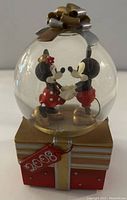 Front view of the snow globe showing Mickey and Minnie Mouse holding hands inside the clear globe on a gift box base with golden bow and 2008 red tag