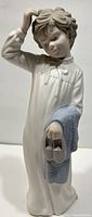 Front and slightly angled view of the NAO by Lladró matte finish figurine showing the boy holding slippers and a blanket, and scratching his head.