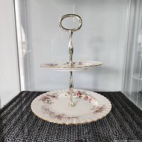 Two-tiered cookie tray with floral clusters and gold trim, displayed on a black woven mat, showing the side profile and tiered structure