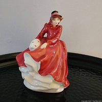 Photo of the Royal Doulton bone china figurine showing full figure of woman seated in red coat and hat holding a white mask.