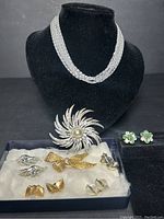 Photo showing the full lot including the silver tone 7 strand choker on a black stand, the large sunburst brooch, and a box containing multiple pairs of clip-on earrings and brooches, and the green and clear gemstone earrings set on black surface.