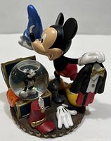 Front and side angle showing Mickey holding wizard hat with tuxedo jacket in other hand and treasure chest with snow globe.