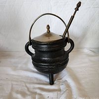 Front and side view of black cast iron cauldron with brass lid, three short legs, two handles, and a metal carrying handle over the top. Pumice stone wand placed through hole in brass lid.