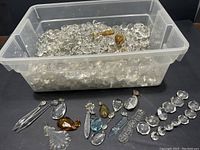 Plastic bin filled with mostly clear and some amber-colored crystal chandelier prisms, with several different prism shapes displayed in front of the bin.