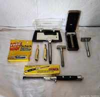 Vintage metal Shick injector razor, two cartridges, sealed cartridge box, two smaller cartridges, and folding pocket comb in box displayed together on a light fabric background.