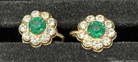 Close up photo showing the front view of a pair of round flower pattern screw back earrings with emerald green center stone and clear rhinestones around it in gold-tone setting.