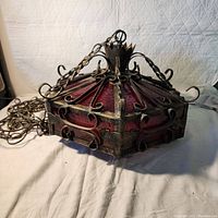 Wrought iron swag light with red stained glass panels and original chain, shown outdoors from side angle to display shape and detail.