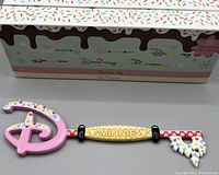 Collectors key with a pink Disney 'D' shape, gold 'MINNIE' plaque, red and white polka dot shaft and multi-colored heart decorations. Box shows dessert themed graphics and other keys in the series.