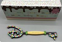Close-up of the Disney Pluto Foodie Series collectible key showing the decorative handle, Pluto label, and key blade with candy-themed packaging in the background.