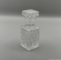 Clear vintage glass perfume bottle with square stopper and geometric embossed pattern on all sides. Photo taken from side angle with neutral background.