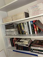 Three shelves showing a collection of cookbooks, stacked cooking magazines, and various fiction and reference books, all on white shelves.