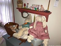 Wide shot showing stuffed bunny dolls, large teddy bear in sweater, baby doll in wooden doll crib, red wall shelf with figurines and vintage boxes.