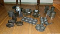 Wide shot of all items showing flat irons, bundt pans, muffin tins, and metal graters on wooden floor.