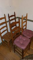 Four wooden ladder-back chairs with woven seats, two topped with red patterned padded cushions, arranged against a wall on hardwood floor.