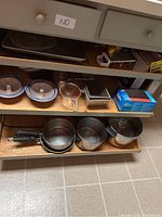 Photo of shelves displaying various kitchen cookware items including saucepans, sauté pan, ceramic bowls with lids, measuring cups, loaf pans, and a colander.