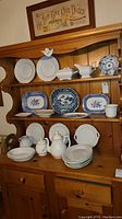 Full view of wooden shelf holding a variety of white and blue and white patterned dishware and servingware, including plates, bowls, a covered dish, gravy boat, and mugs.