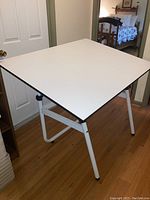 White adjustable drafting table shown angled, with metal legs and flat surface