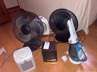 Photo of two black oscillating fans, one white fixed fan, two tabletop heaters (white cylindrical and black cube-shaped), and a Black and Decker handheld vacuum cleaner on wooden floor.